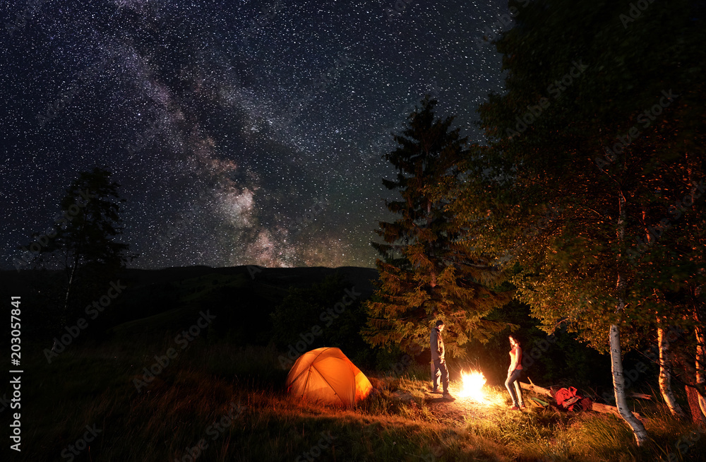 Naklejka Guy and girl backpackers standing by the campfire near the woods and illuminated orange tent under the starry sky which is clearly visible milky way. Night camping near forest in the mountains