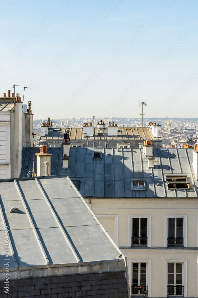 Fototapeta premium Open view over the rooftops of Montmartre district in Paris with typical zinc roofing, skylights, chimneys and TV antennas under a clear blue sky.