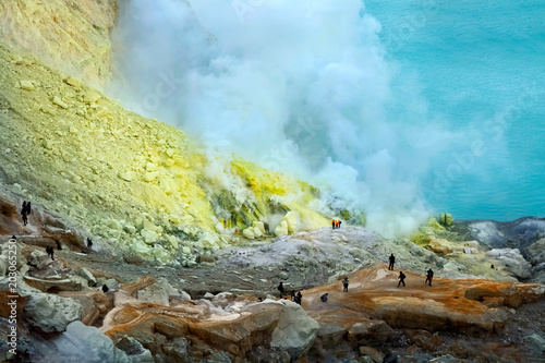 Small silhouettes of people in the crater of the Ijen volcano against the background of yellow sulfuric rocks and a sulphurous blue lake. Volcanic caldera of an active volcano. Indonesia.