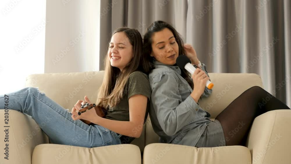 Happy and positive sisters playing and singing on the sofa in the living room. The youngest is playing at ukulele while the oldest is singing in a microphone. They are sitting on the couch with their Happy and positive sisters playing and singing on the sofa in the living room. The youngest is playing at ukulele while the oldest is singing in a microphone. They are sitting on the couch with their