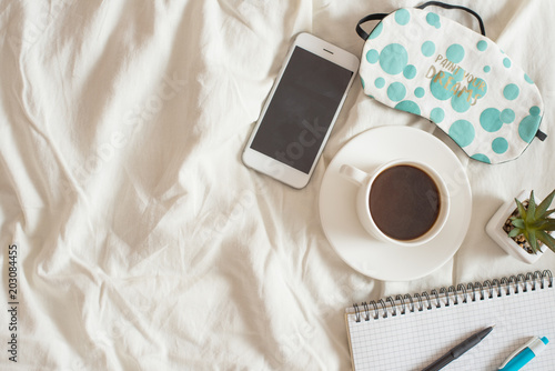 A cup of coffee, with an alarm clock, a phone, and a notebook, on a brown plaid on the bed. The concept of the morning. Top view with empty space for inscription