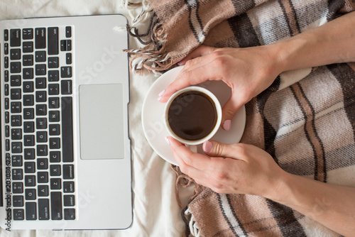 A cup of coffee in the hands of a girl and a laptop on a brown plaid. Top view, morning concept