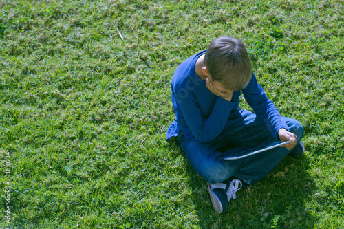 Boy with tablet computer sitting on the grass at sunny day outdoors.