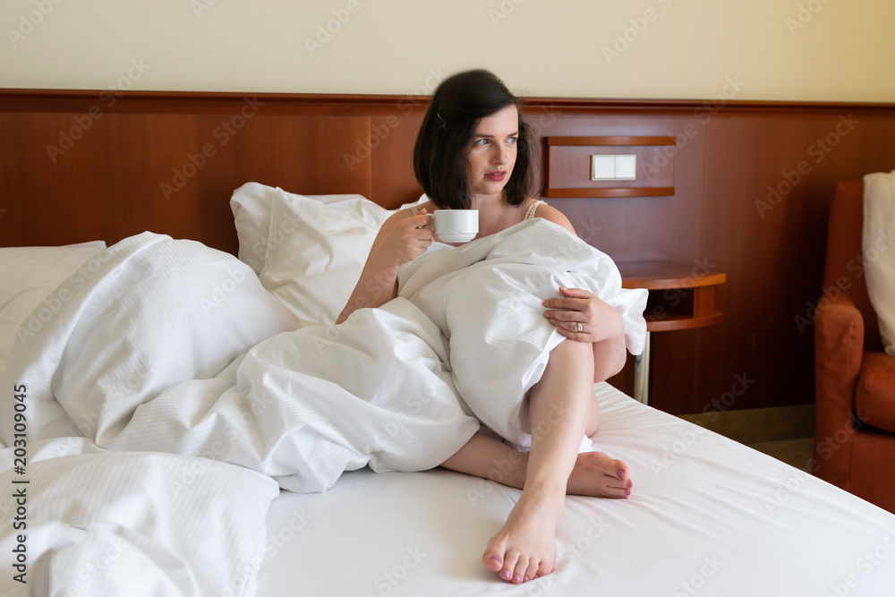 An attractive young caucasian brunette woman drinking coffee in bed in hotel room An attractive young caucasian brunette woman drinking coffee in bed in hotel room