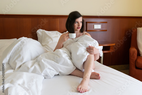 An attractive young caucasian brunette woman drinking coffee in bed in hotel room