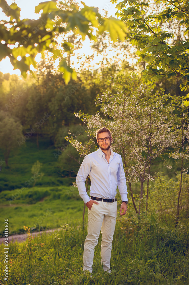Portrait of trendy handsome hipster man in eye glasses posing in spring park alone at sunset ao apple tree nature background. Young businessman at vacation
