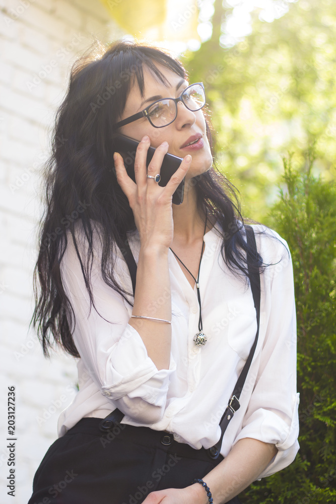 Beautiful business woman talking on the phone. A successful female with glasses is holding a smartphone in her hand on a summer street. Beautiful business woman talking on the phone. A successful female with glasses is holding a smartphone in her hand on a summer street.