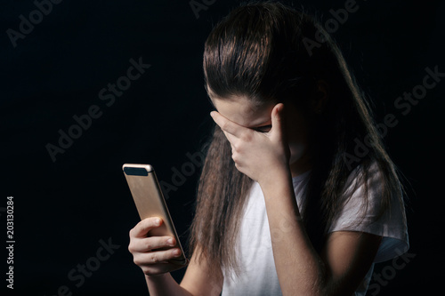 Single sad teen holding a mobile phone lamenting sitting on the bed in her bedroom with a dark light in the background photo