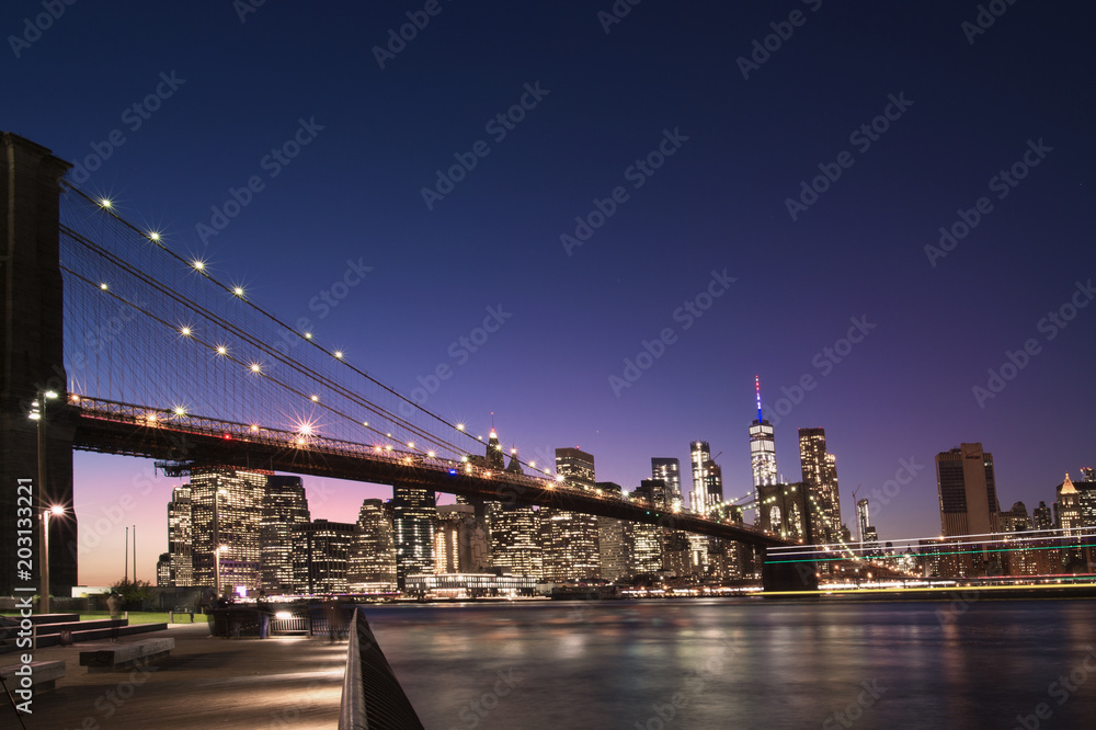 Fototapeta premium Brooklyn Bridge and the illuminated Skyline of Manhattan in the evening with blue sky and smooth water surface shot from Brooklyn side, New York, Usa.