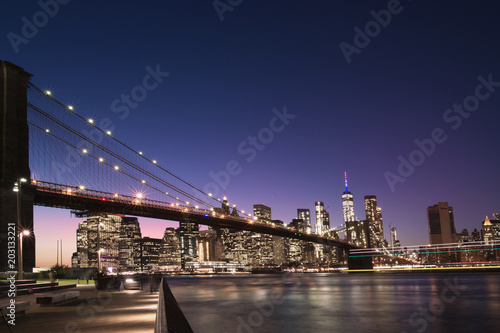 Fototapeta Naklejka Na Ścianę i Meble -  Brooklyn Bridge and the illuminated Skyline of Manhattan in the evening with blue sky and smooth water surface shot from Brooklyn side, New York, Usa.