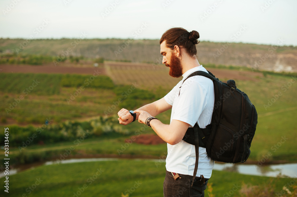 Fototapeta premium Bearded hipster tourist man touching his smartwatch standing in nature at sunset