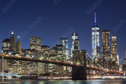 Brooklyn Bridge and the illuminated Skyline of Manhattan in the evening with blue sky and smooth water surface shot from Brooklyn side, New York, Usa.