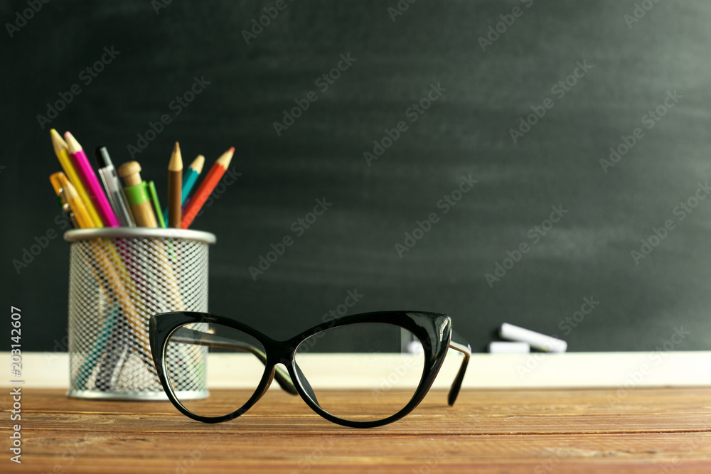 Glasses teacher and a stand with pencils on the table, on the background of a blackboard with chalk. The concept of the teacher's day. Copy space. Glasses teacher and a stand with pencils on the table, on the background of a blackboard with chalk. The concept of the teacher's day. Copy space.