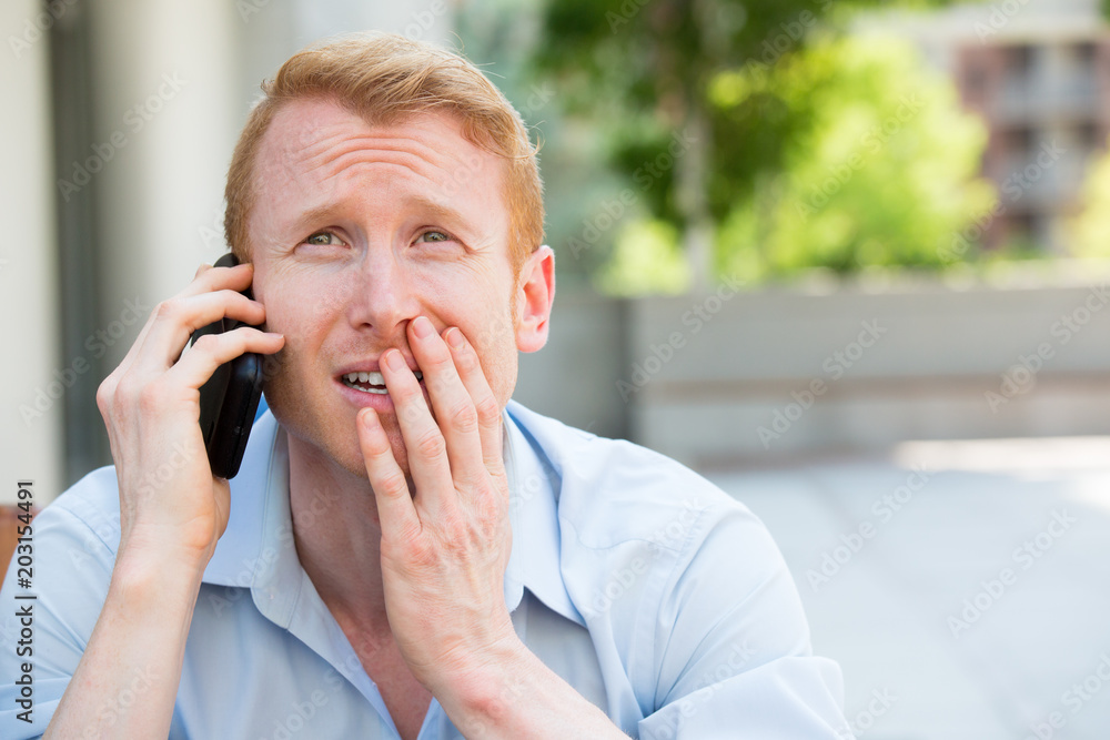Closeup portrait, worried young man in blue shirt talking on phone to someone, looking gloomy, isolated outdoors outside background Closeup portrait, worried young man in blue shirt talking on phone to someone, looking gloomy, isolated outdoors outside background