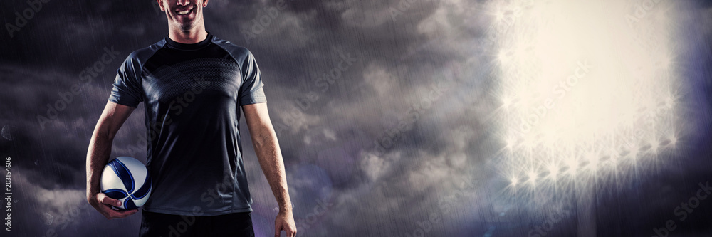 Full length portrait of smiling rugby player in black jersey holding ball against spotlight in the sky Full length portrait of smiling rugby player in black jersey holding ball against spotlight in the sky