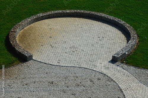 Small round floor tiled plateau surrounded by stones in steel cages, also called gabions in center of cultivated lawn. Crushed stone is surrounding stone tiled pathway photo