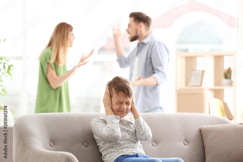 Little unhappy boy sitting on sofa while parents arguing at home Stock
