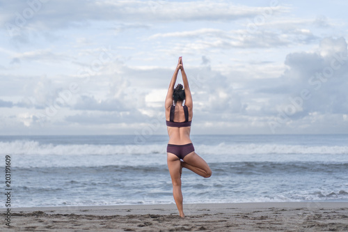 rear view of woman practicing yoga in tree pose (Vrksasana) on seashore