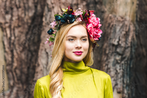 smiling young woman with floral wreath looking away with blurred tree trunk on background