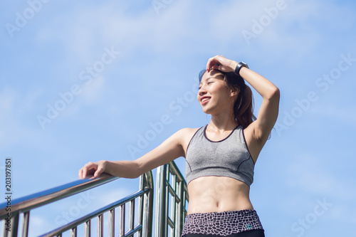 Young woman relaxing after jogging exercise on fence at park to freshen her body and enjoy warm light in morning. Young asian woman take a break from running execise. Outdoor exercise activity. © tirachard