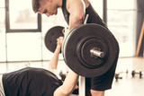 handsome trainer helping sportsman lifting barbell with heavy weights in gym