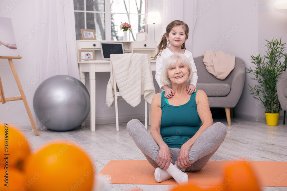 Fototapeta premium Happiness. Cheerful old grandmother sitting on the carpet and her granddaughter hugging her from behind
