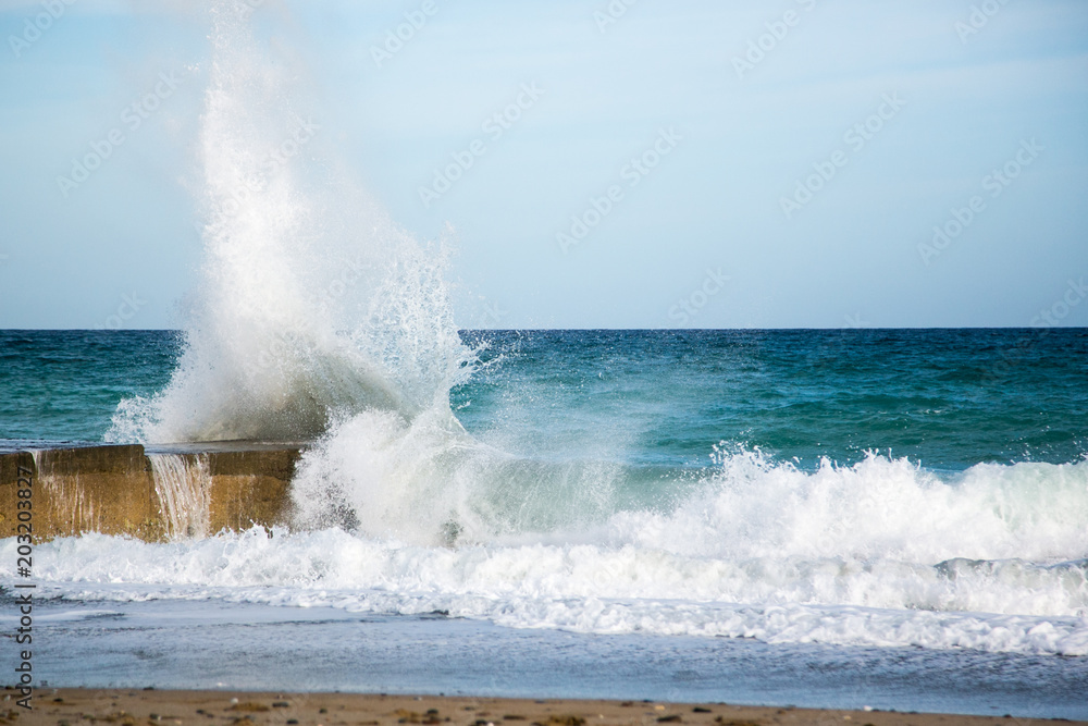 Fototapeta premium Wasserkraft. Eine Welle klatscht mit voller Wucht gegen einen Felsen an der Küste Kretas, im Vordergrund Sandstrand und Brandung, im Hintergrund das Mittelmeer und blauer Himmel .