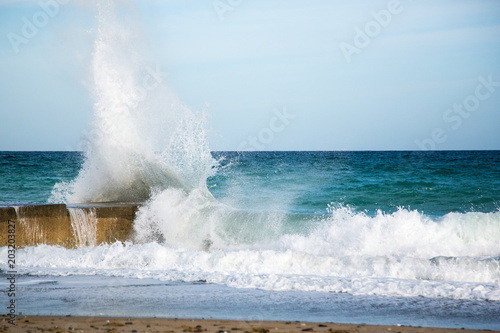 Wasserkraft. Eine Welle klatscht mit voller Wucht gegen einen Felsen an der Küste Kretas, im Vordergrund Sandstrand und Brandung, im Hintergrund das Mittelmeer und blauer Himmel .