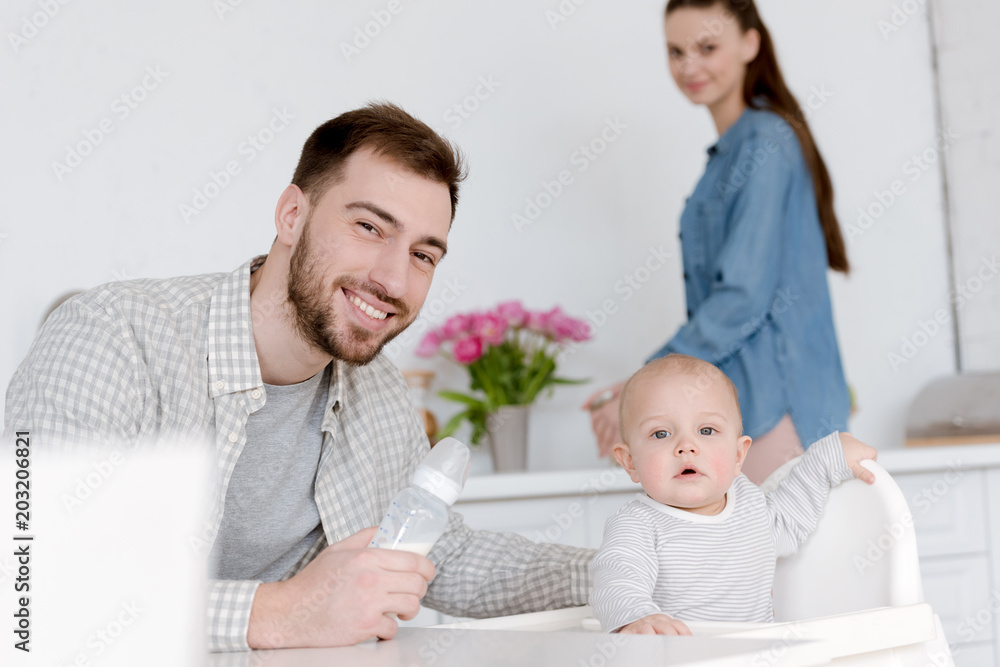 father feeding son with milk in baby bottle, mother standing on kitchen behind father feeding son with milk in baby bottle, mother standing on kitchen behind