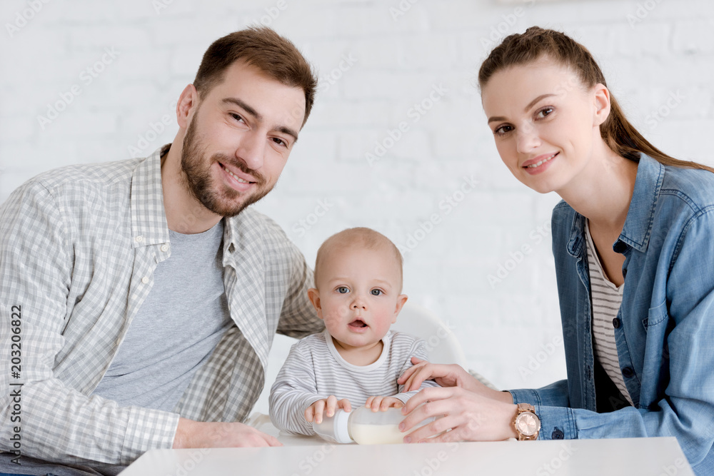 smiling parents with little son sitting on kitchen with baby bottle smiling parents with little son sitting on kitchen with baby bottle