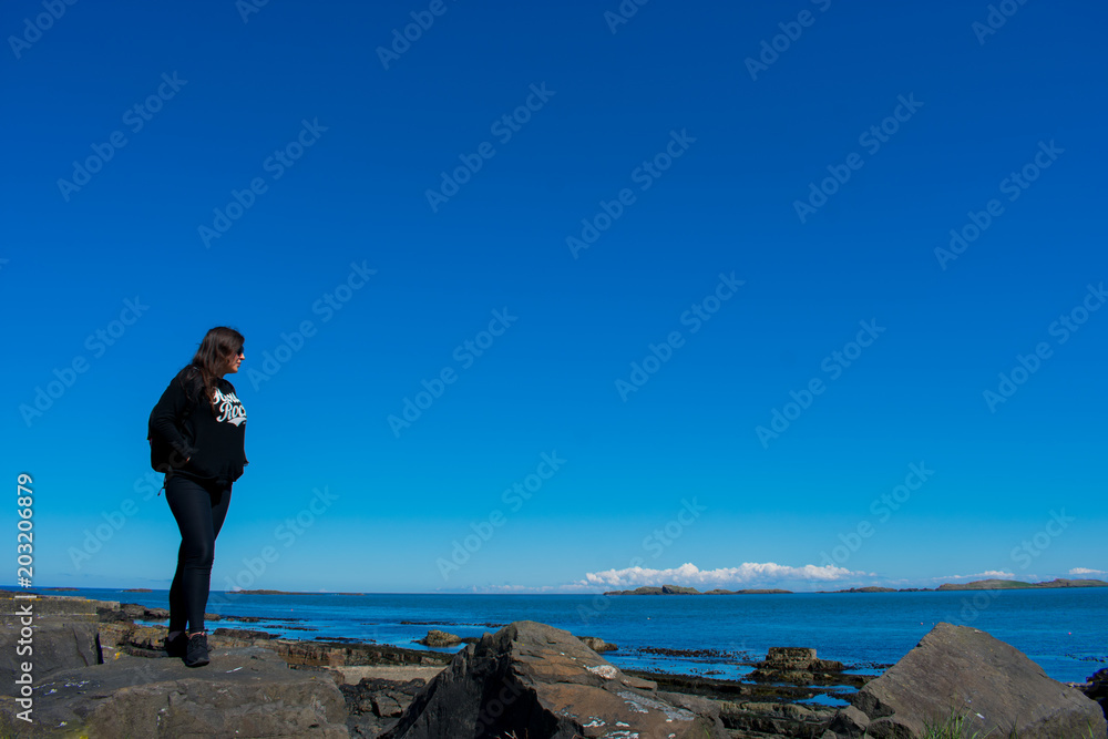beautiful woman with dark hair dressed black standing on rock formations and looking on ocean. thinking about live 