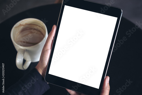 Top view mockup image of hands holding black tablet pc with white blank screen and coffee cup on table background