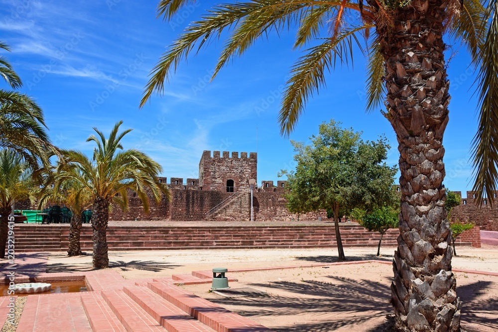 Palm trees and landscaped gardens within the Medieval castle with battlements and tower to the rear, Silves, Portugal.