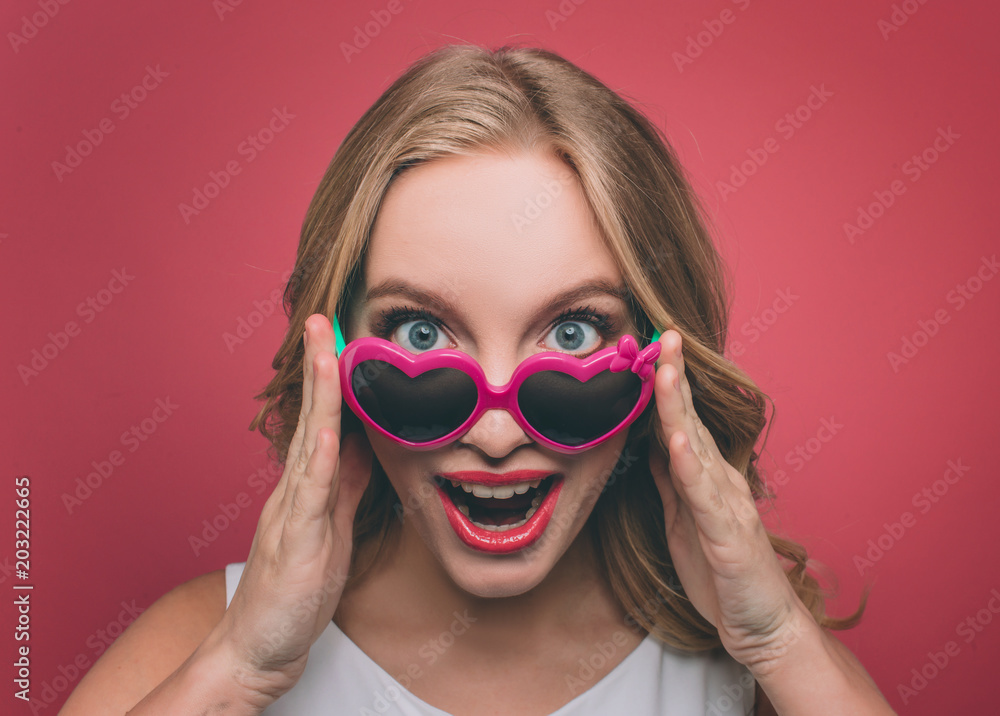 Pretty woman with blonde hair weard sunglasses with pink edge. She is looking on camera and holding glasses at the eage of it. She happy and good-looking. Isolated on pink background.