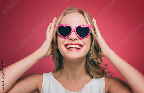Beautiful girl wears glasses with pink edge. She is looking up and smiling. Girl is holding her handson the head. Isolated on pink background.