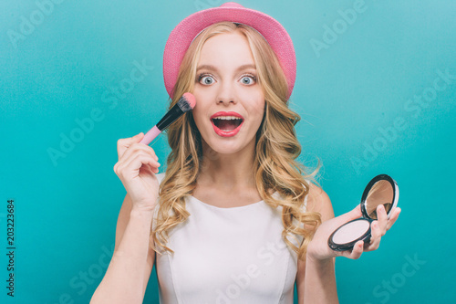 Happy girl with blonde hair wears pink heat. She has some cosmatics in her hands. Girl is putting some blush on her chicks with a brush. In other hand she has some blush. Isolated on blue background. photo
