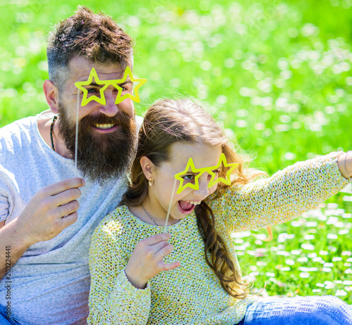 Dad and daughter sits on grass at grassplot, green background. Child and father posing with star shaped eyeglases photo booth attribute at meadow. Star and fame concept. Family spend leisure outdoors. photo