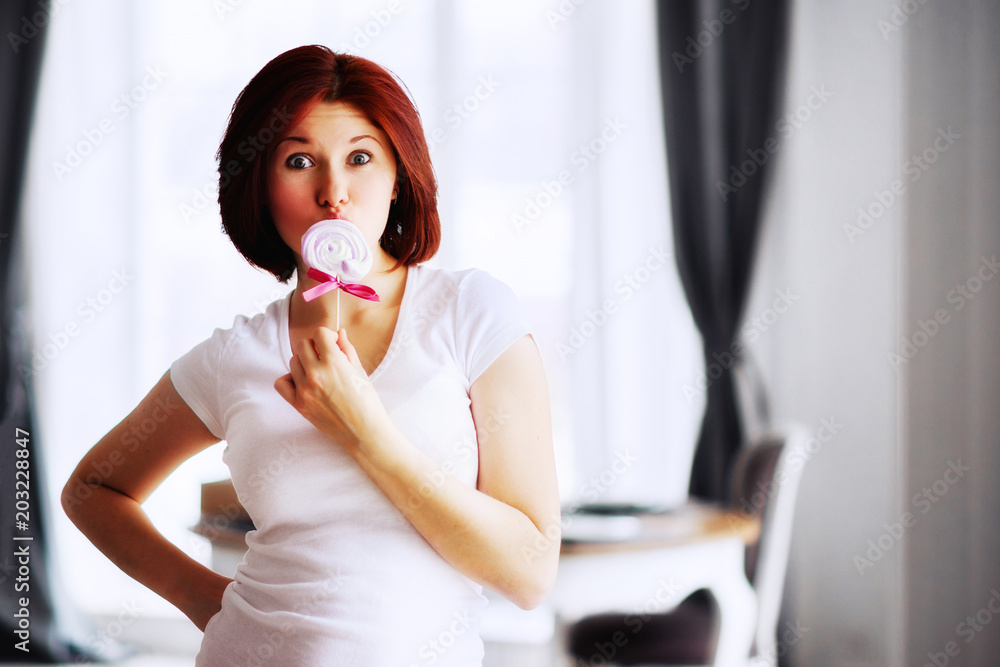 Portrait of young red headed woman with a candy on stick. Beautiful woman standing with surprised face covering her lips with candy. Portrait of young red headed woman with a candy on stick. Beautiful woman standing with surprised face covering her lips with candy.