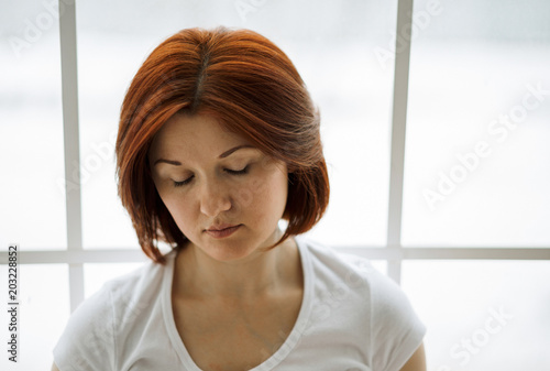 Young depressed woman with closed eyes. Portrait of an upset and devastated woman sitting with her back to window. Sad emotions, bad feelings and depression concept