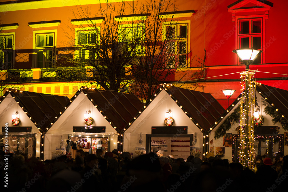 Crowded Christmas market in city center, decorated wooden huts with glowing lights. People enjoying event and searching for gifts. Beautiful Christmas background