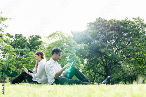 Young asian couple,lover holding earphone,smartphone sit relax in park,tree background copy space. Happy outdoor on summer,spring sunshine in city green park,garden lifestyle. Take a break after work.