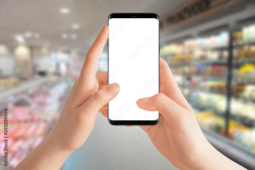 Female hands holding modern smartphone with isolated screen, blurred grocery store in background. Online shopping concept, Mockup