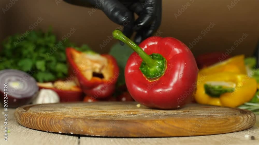 Chef with black gloves, lays juicy red pepper with flowing droplets of water vracha him and examining on all sides on a wooden board on a background of greenery, red pepper, brocoli, background blur Chef with black gloves, lays juicy red pepper with flowing droplets of water vracha him and examining on all sides on a wooden board on a background of greenery, red pepper, brocoli, background blur