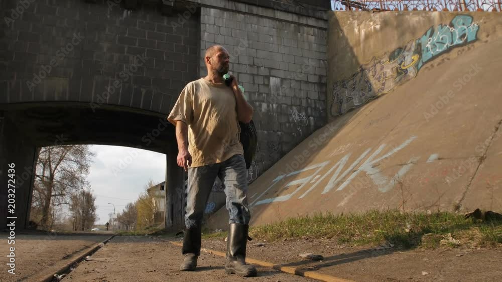 Front view full length of mature bearded homeless man in dirty clothes walking near bridge tunnel in city. Male in boots holding plastic bin bags with trash and belongings. Steadicam stabilized shot. Front view full length of mature bearded homeless man in dirty clothes walking near bridge tunnel in city. Male in boots holding plastic bin bags with trash and belongings. Steadicam stabilized shot.