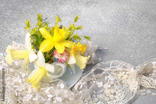 Fototapeta Naklejka Na Ścianę i Meble -  yellow narcissus and wild spring flowers bouquet in a coffee cup instead of vase with lace and copy space for your greetings text