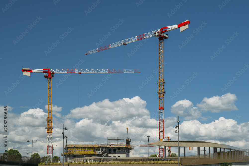 Construction cranes and unidentifiable construction workers at a building site with blue sky and copy space. Construction cranes and unidentifiable construction workers at a building site with blue sky and copy space.