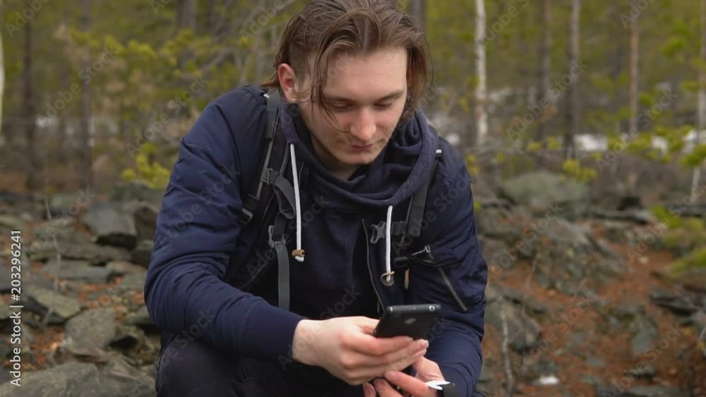 A young tourist with a backpack on his back pokes his finger in the screen of the mobile phone. In the background there is a coniferous forest with stony soil. A young tourist with a backpack on his back pokes his finger in the screen of the mobile phone. In the background there is a coniferous forest with stony soil.