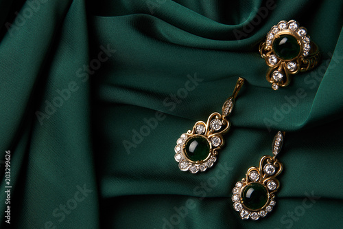 Beautiful Golden ring and pair of earrings with green Emerald and Diamonds gemstones on a green satin background. Luxury female jewelry, close-up. Selective focus