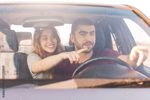 Excited female and male in car, points at windscreen, notice something funny on road, enjoy unforgettable trip in automobile, have good transport for travelling. People and reaching destination