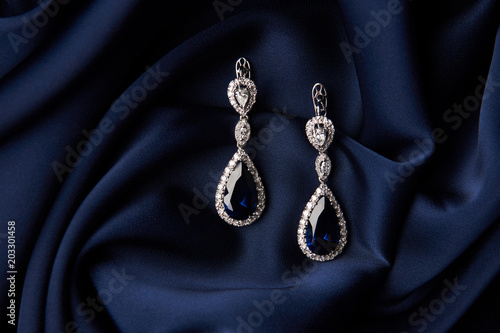 Two Golden sapphire earrings with small diamonds. Pair of platinum earring with sapphire gemstone on blue satin background. Luxury female jewelry, close-up